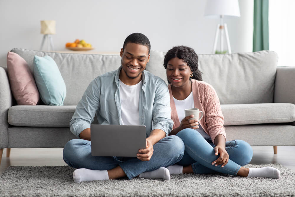 Happy black lovers in casual sitting on carpet by couch in living room
